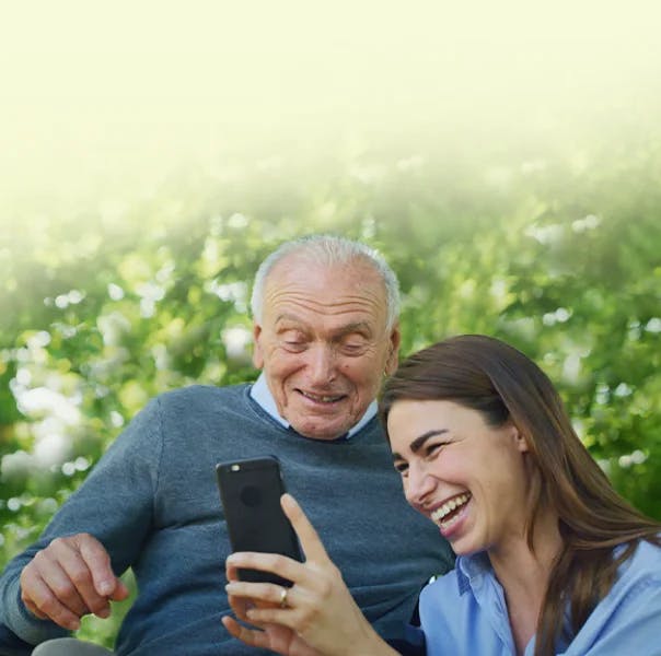 An older man and a young female are looking at a mobile and laughing.